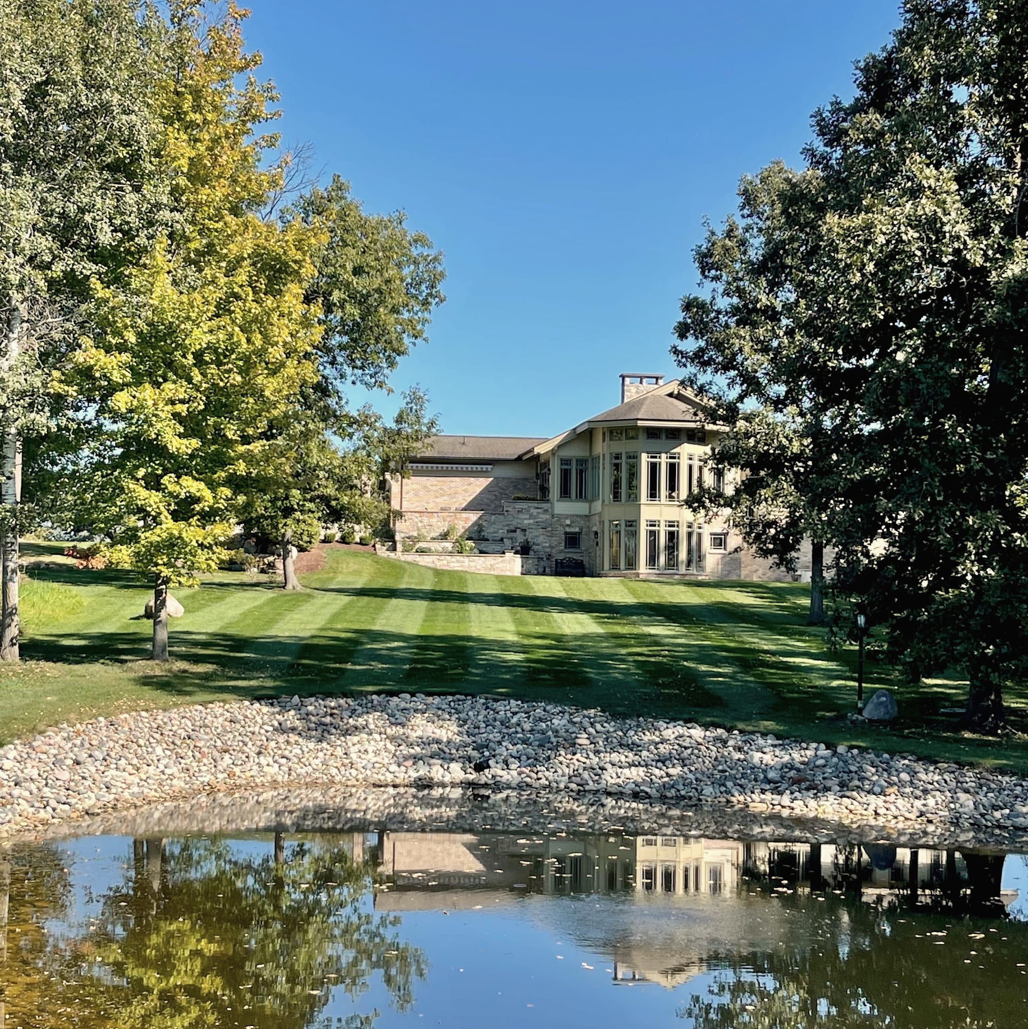 A striped lawn sloping down to a pond with a stone edge and a large house behind trees