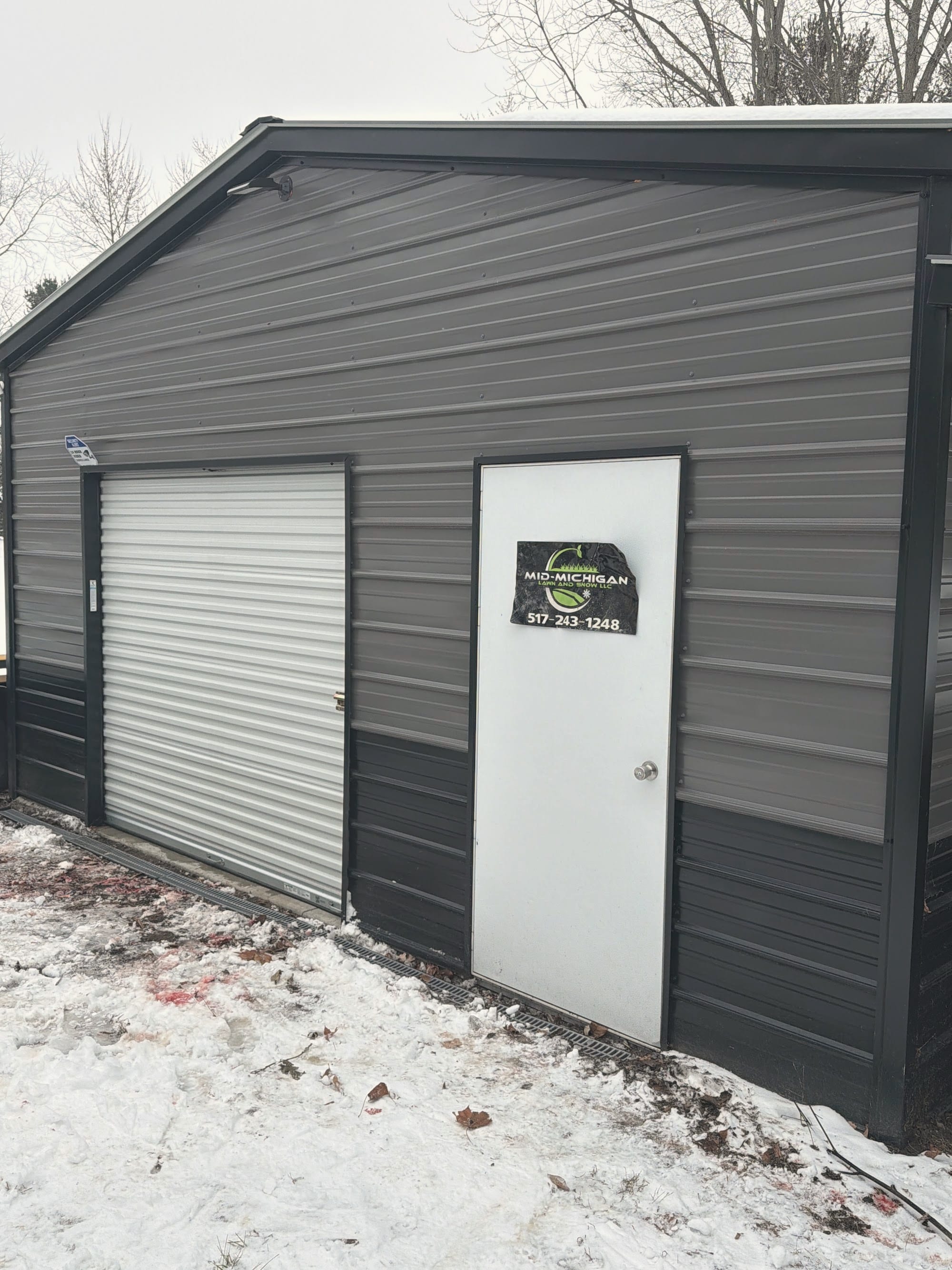 A gray metal shop building with a Mid-Michigan Lawn and Snow sign on the door, snow on the ground